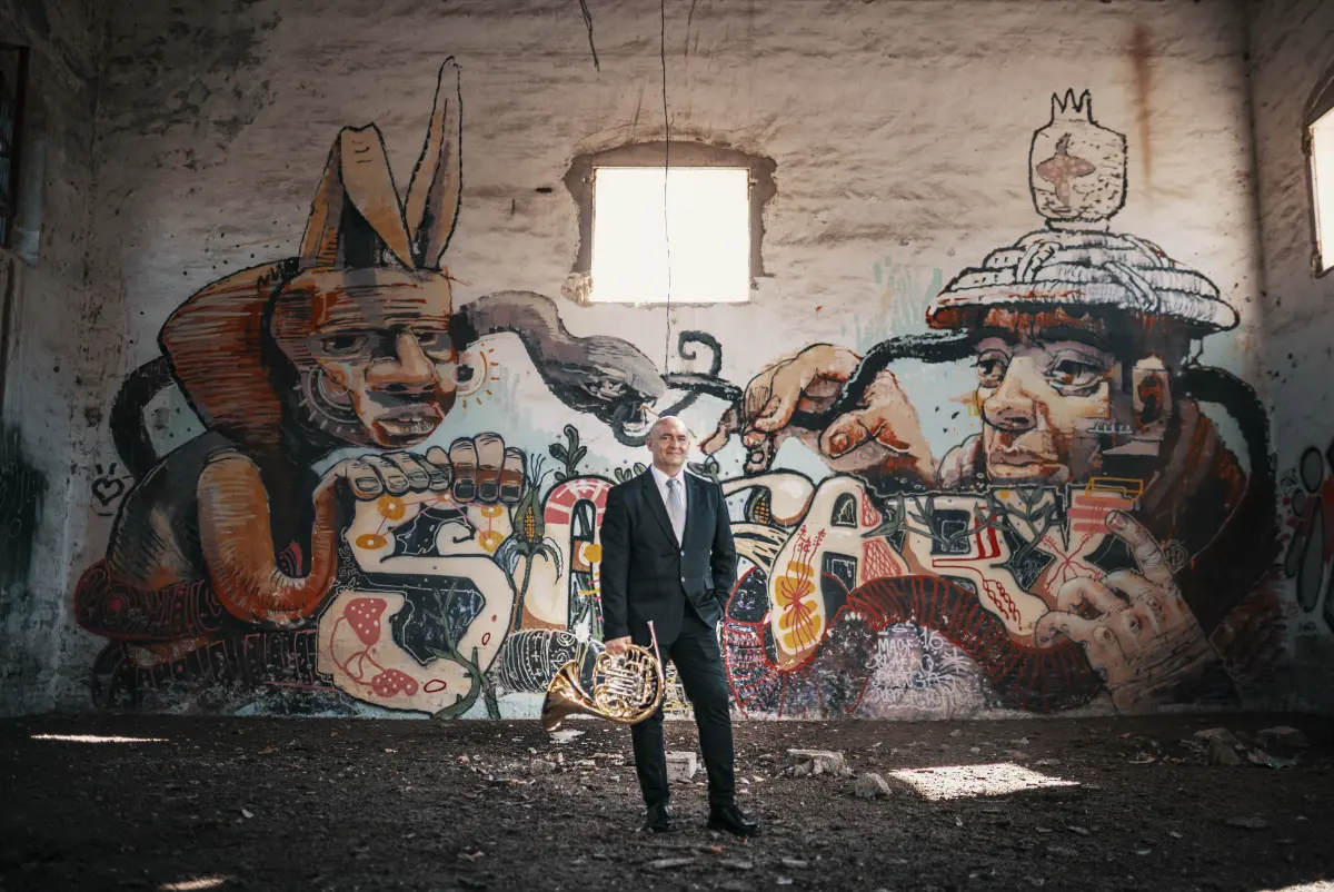 Oscar Sala de pie con su trompa ante un gran mural, en una nave industrial
